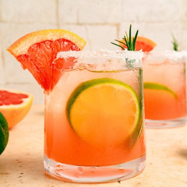 Side view of a paloma cocktail on a kitchen counter, garnished with citrus and rosemary.