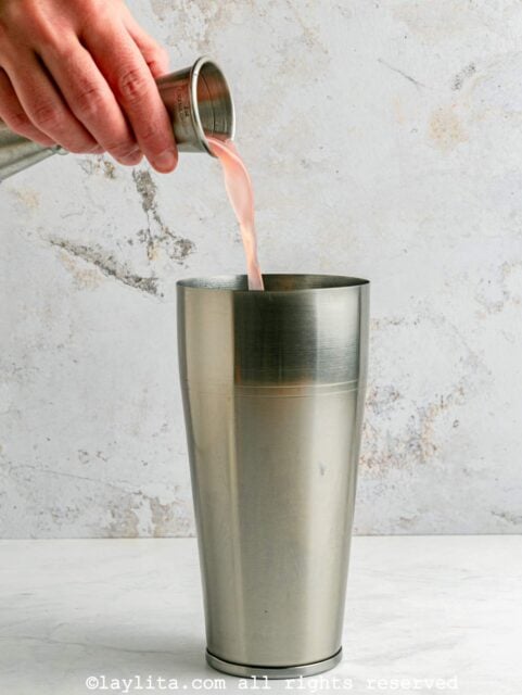 Preparing a paloma cocktail in a shaker, with a hand pouring in a measure of grapefruit juice