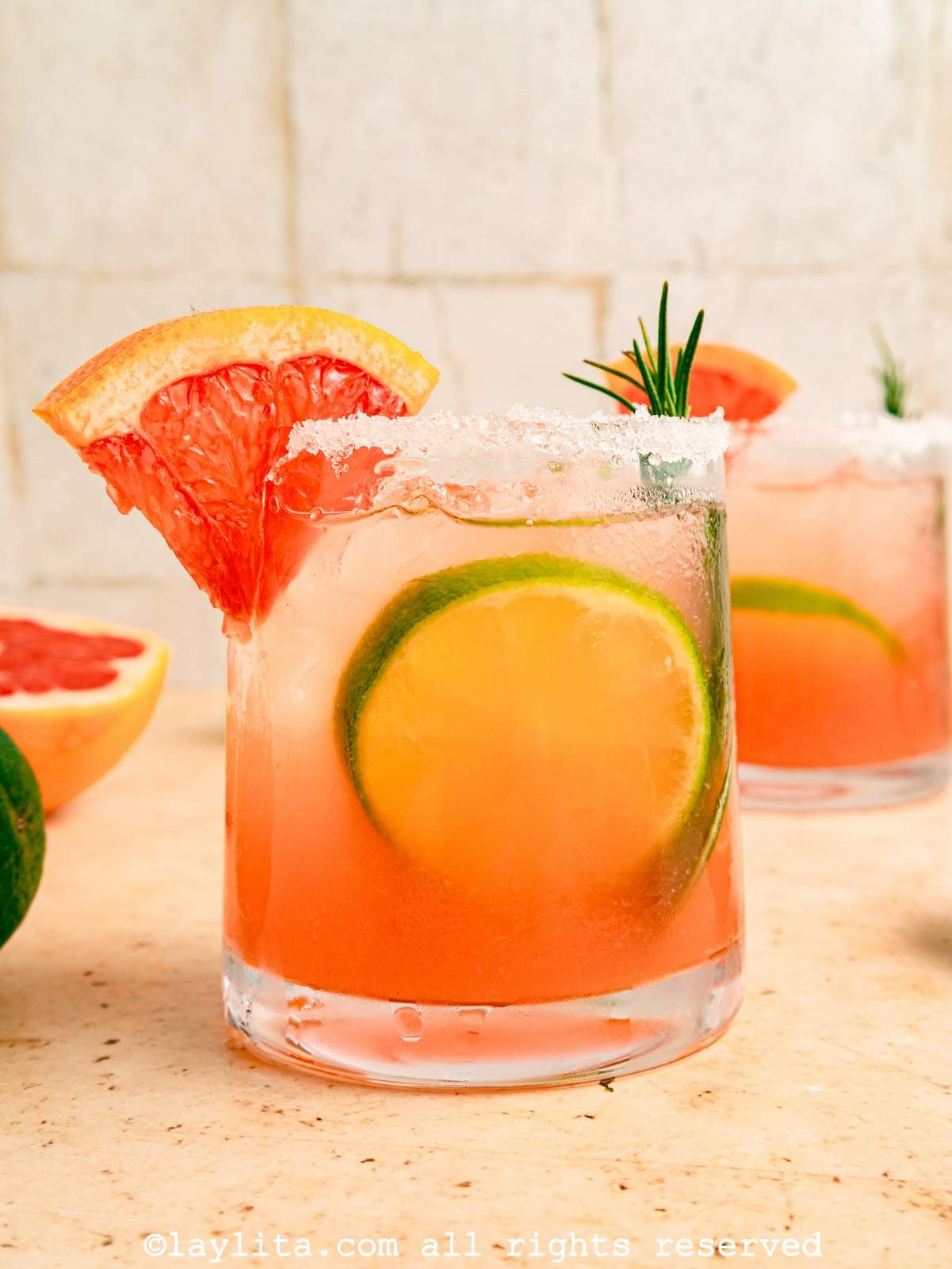 Side view of a paloma cocktail on a kitchen counter, garnished with citrus and rosemary.
