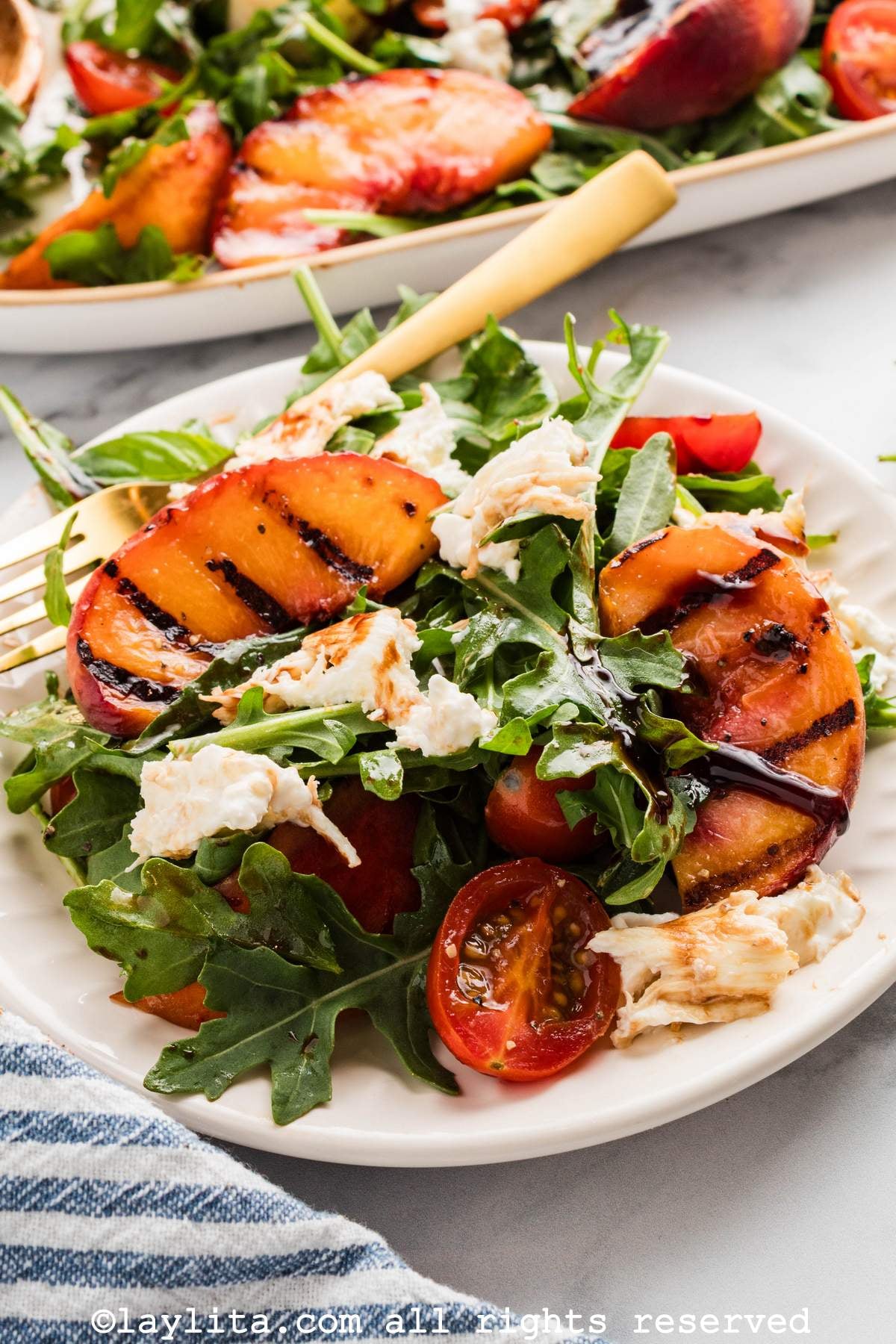 Grilled peach salad with arugula and pieces of burrata cheese on a plate, with more salad in the background