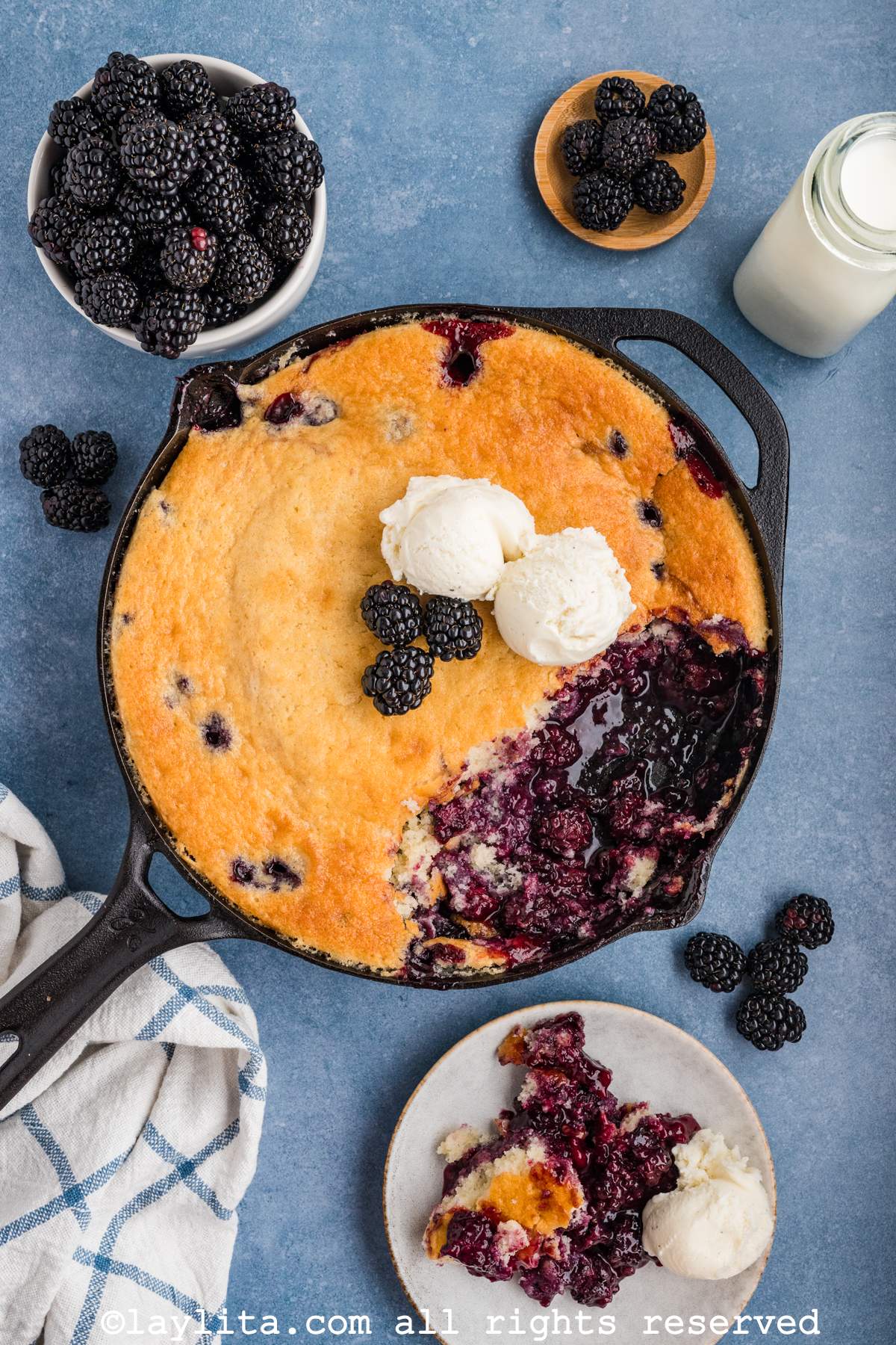 Overhead shot of blackberry cobbler in a cast iron skillet with some missing from the skillet. There are some fresh berries and two scoops of ice cream on top of the cobbler.