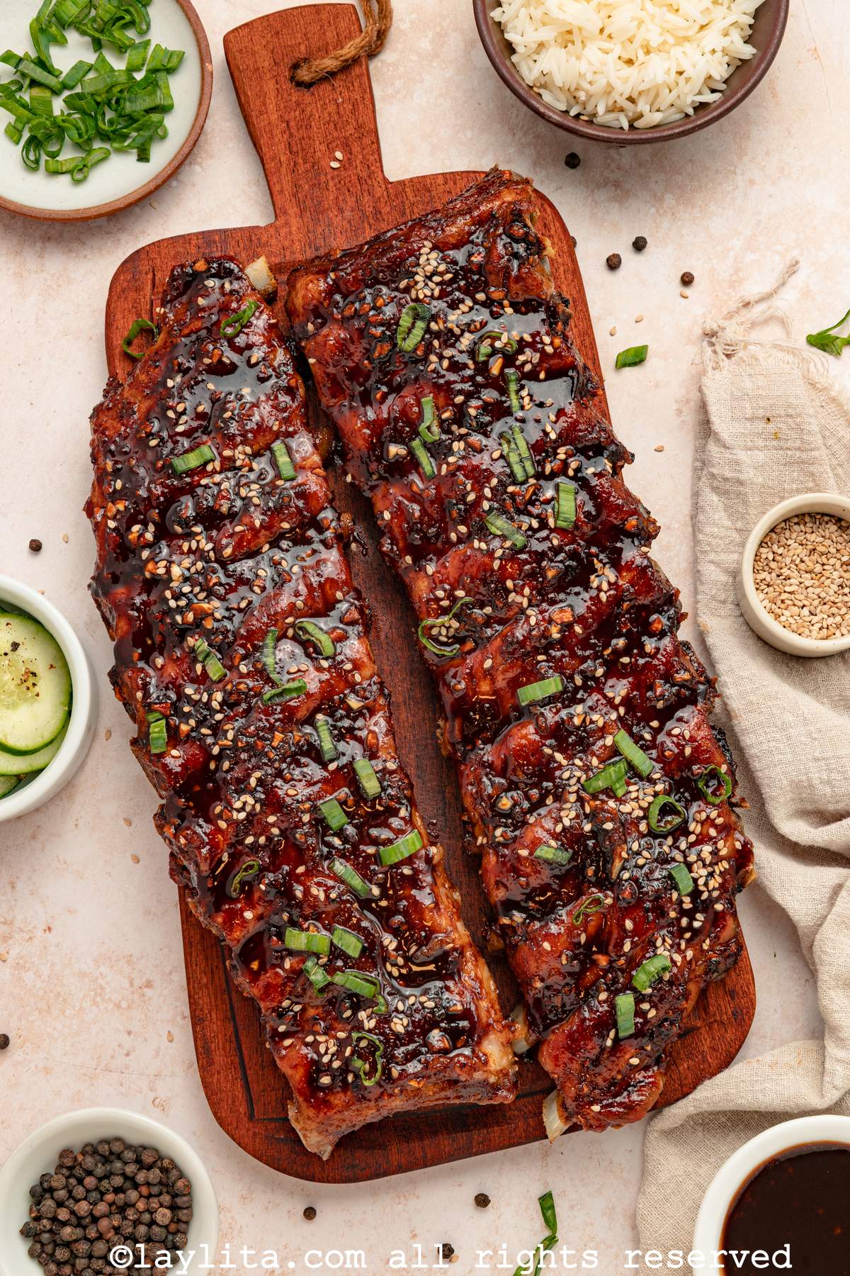 Two racks of Asian sticky ribs on a wood cutting board next to a small bowl of rice, cucumber salad, sesame seeds, herbs and sauce for dipping.