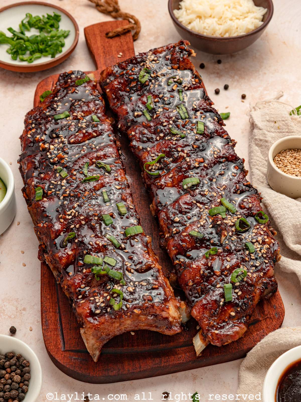 Overhead view of two racks of sticky pork ribs on a wood cutting board next to a small bowl of rice, cucumber salad, sesame seeds, herbs and sauce for dipping.