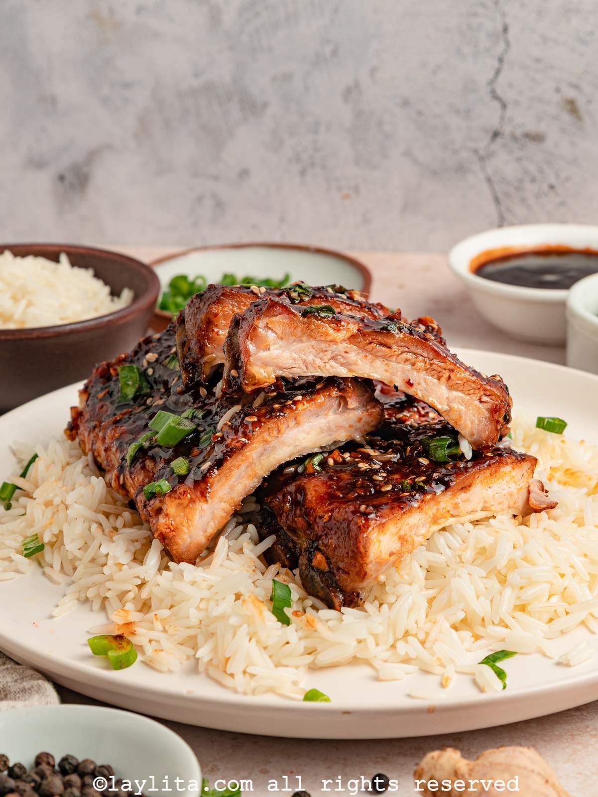 Side view of several sticky pork ribs sliced into pieces and stacked on top of each other in a bowl of rice, with a bowl of sauce and more rice in the background
