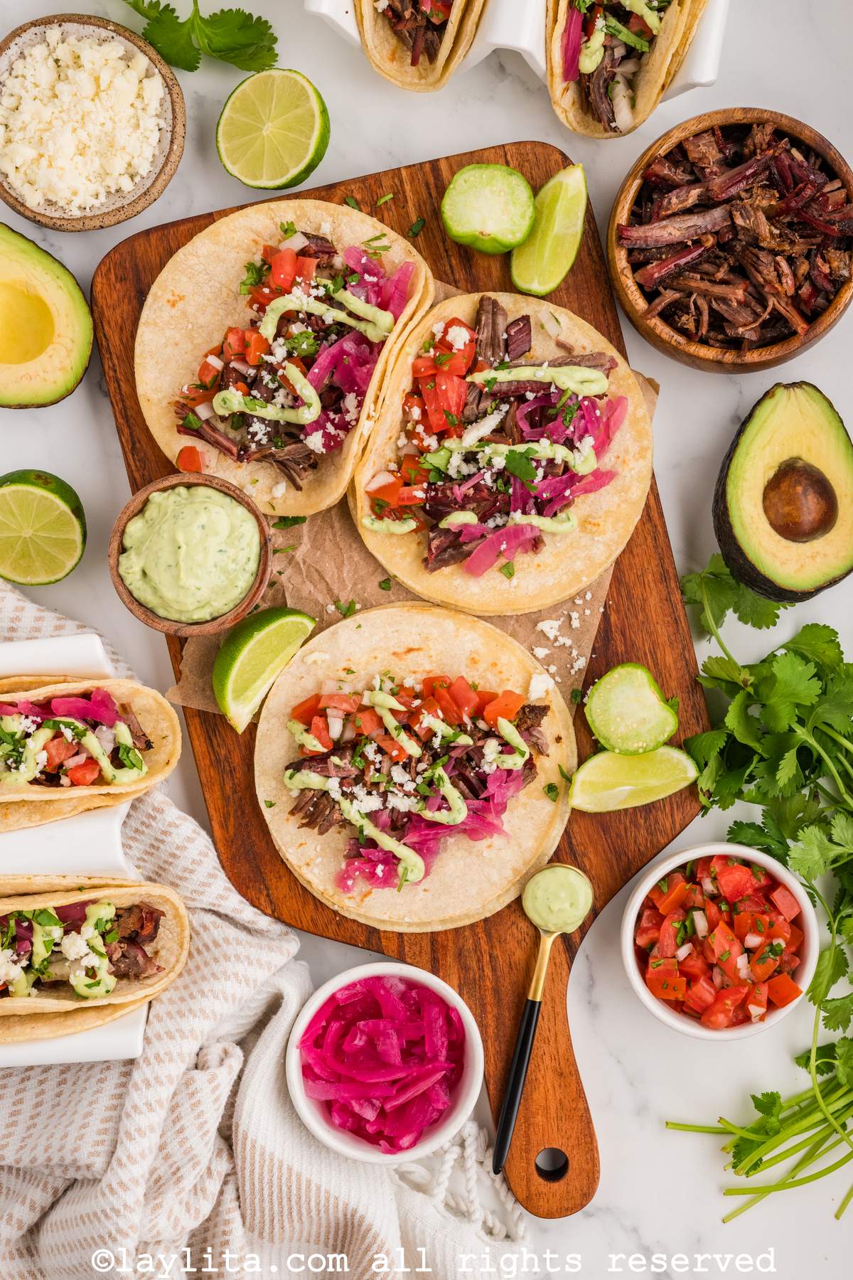 Overhead shot of brisket tacos on a wooden board next to sauces, lime wedges and cilantro.