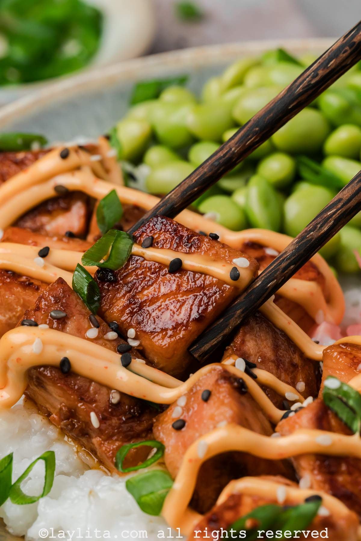 Chopsticks picking up a piece of crispy salmon from a bowl with edamame in the background