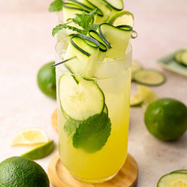 Side view of several glasses lined up and filled with cucumber lime mint drink, garnished with cucumber ribbons, lime slices and mint leaves