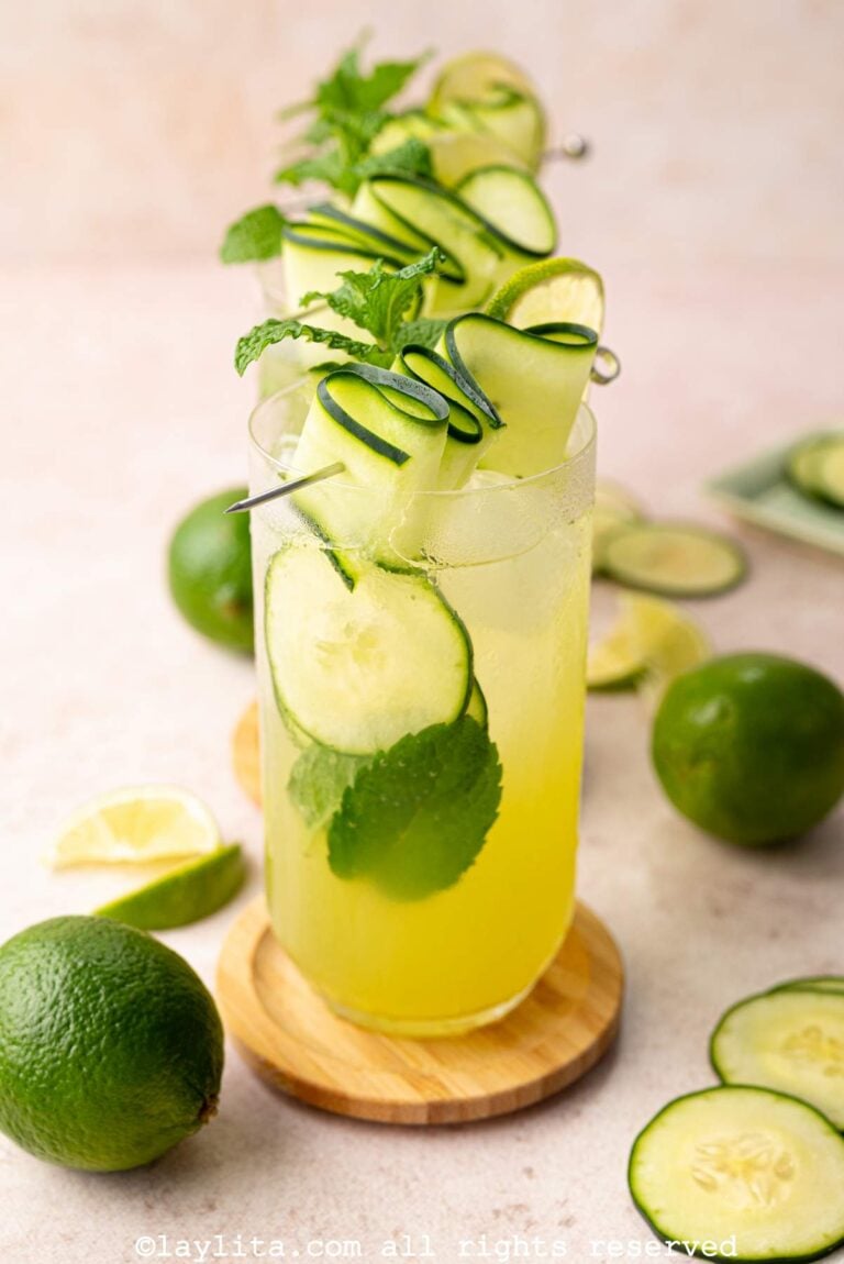 Side view of several glasses lined up and filled with cucumber lime mint drink, garnished with cucumber ribbons, lime slices and mint leaves