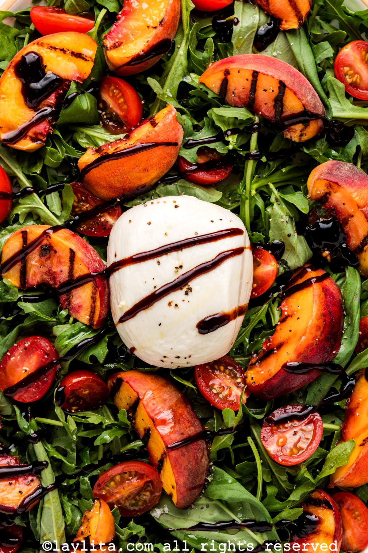 Closeup of a salad with grilled peach slices on top of arugula, with a big ball of burrata cheese in the center and a balsamic glaze drizzle
