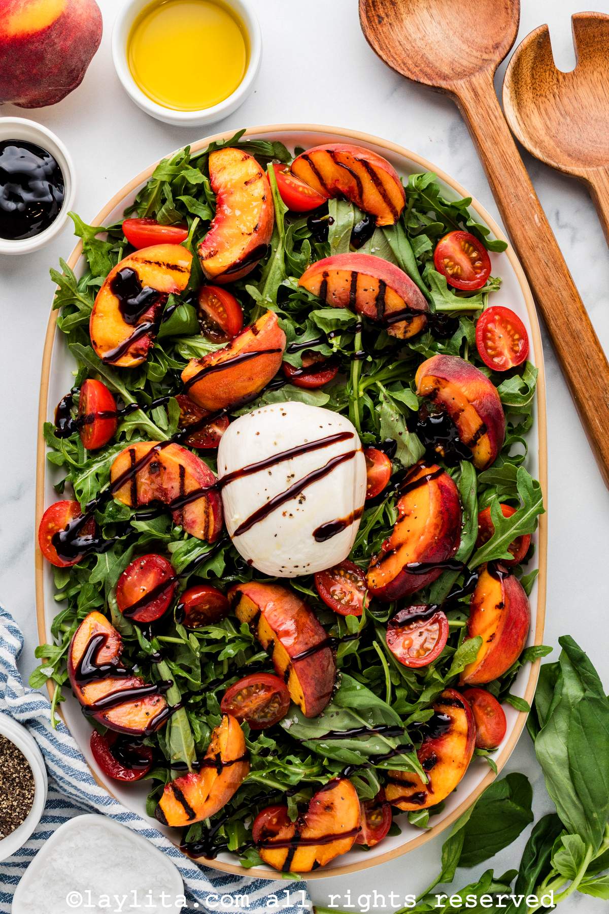 Overhead view of a salad made with grilled peach wedges, cherry tomato, burrata cheese, arugula, basil leaves and balsamic glaze on an oval platter, on the right side there wooden serving utensils and on the left side there is a small bowl of olive oil, some balsamic glaze and a whole peach