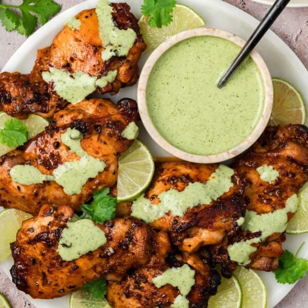Overhead view of a plate of roasted chicken thighs with green sauce drizzled on top, with the bowl of sauce on the late and lime slices on the side