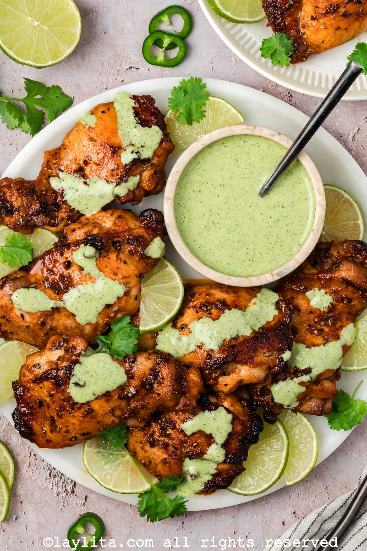 Overhead view of a plate of roasted chicken thighs with green sauce drizzled on top, with the bowl of sauce on the late and lime slices on the side