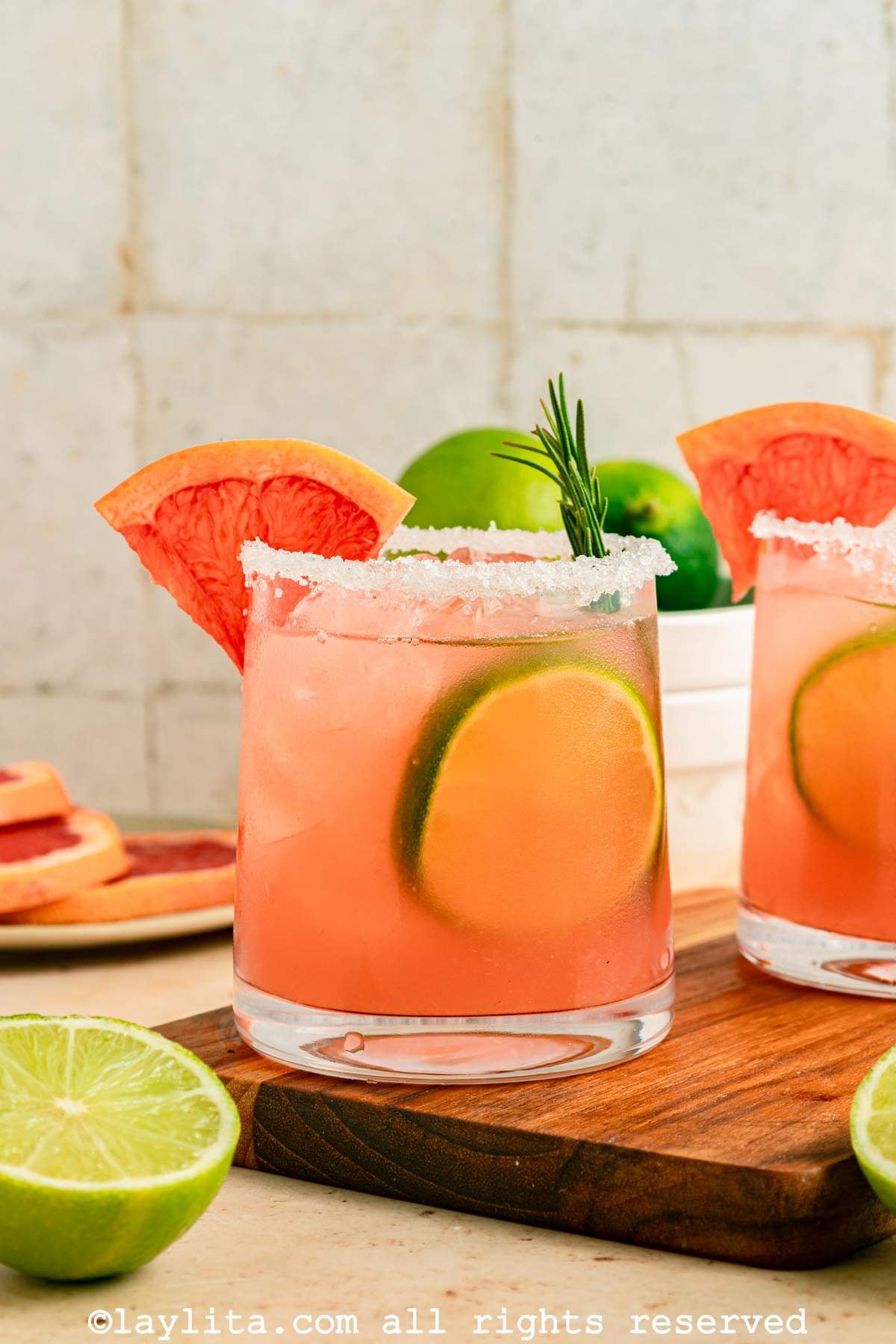 A grapefruit paloma cocktail with salt on the rim, on a wooden board next to a lime, in the background there is a second cocktail glass