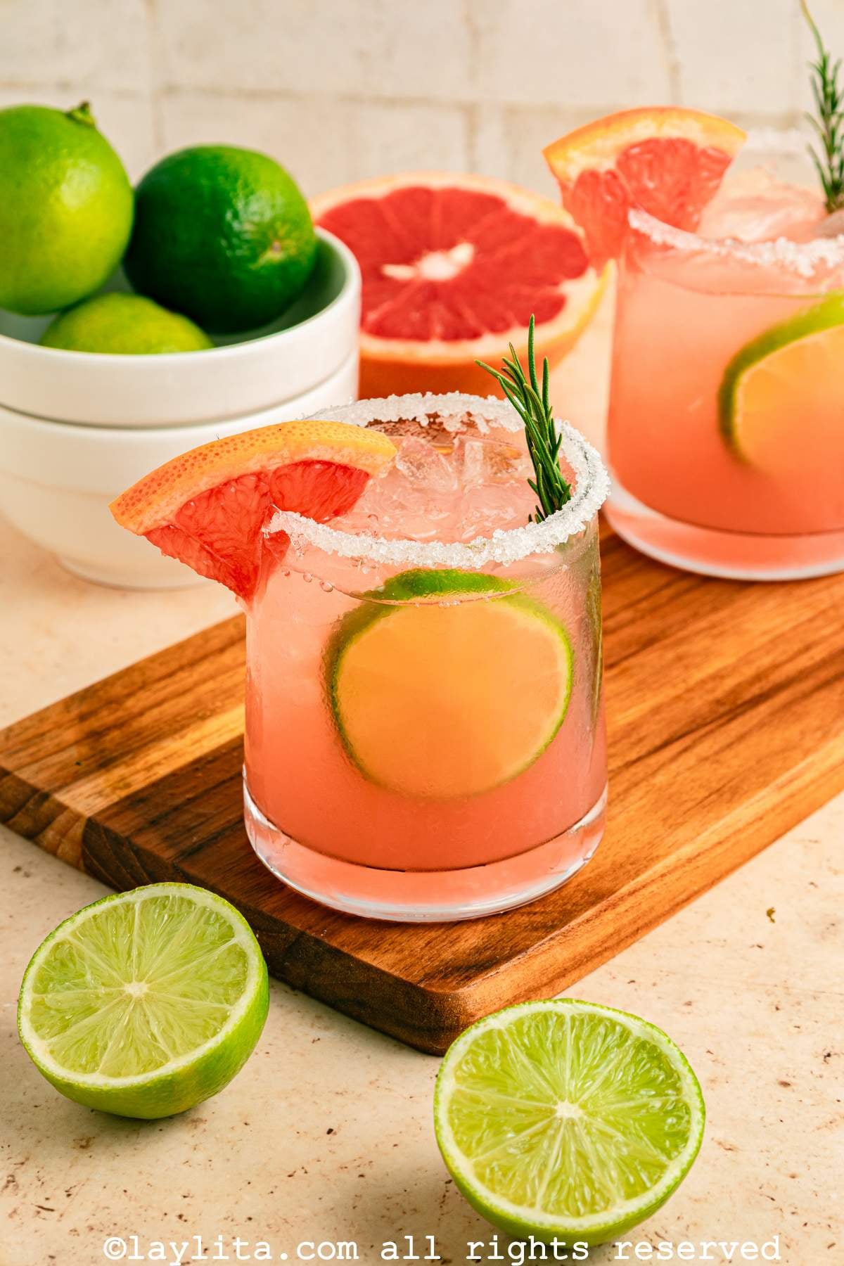 Two paloma cocktails with salt on the rim, on a wooden board next to a lime