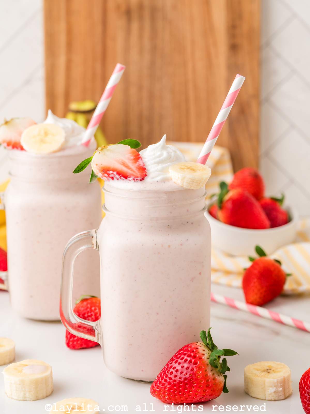 Two strawberry banana milkshakes in large jars, both with straws and garnishes on top.