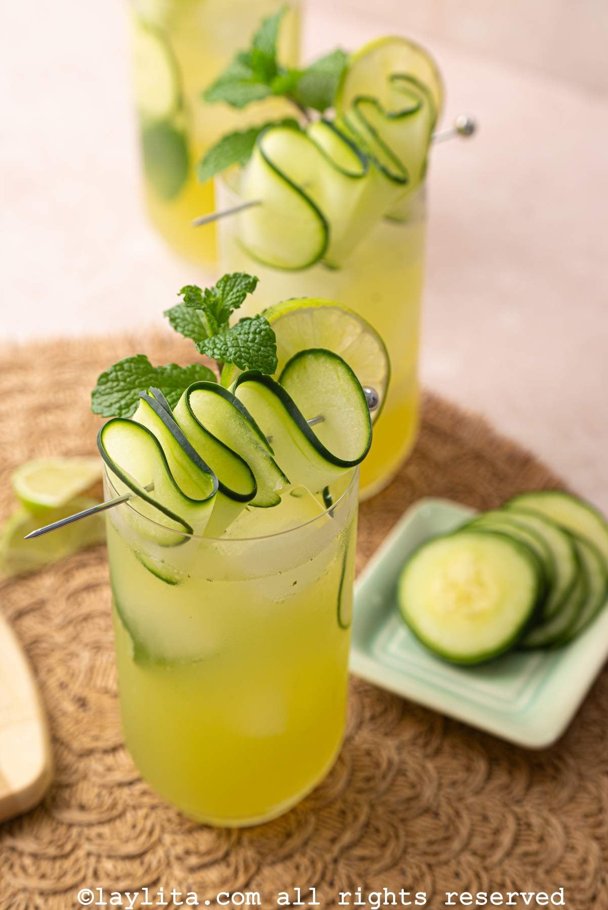 A glass of cucumber lime mint cooler on a kitchen matt, with a plate of cucumber slices on the side and more drinks in the back