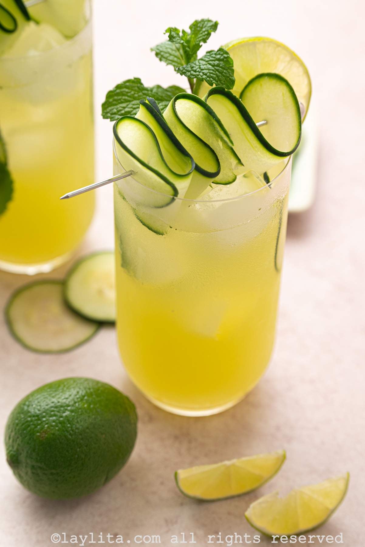 A glass of cucumber lime agua fresca with lime wedges and a whole lime next to it, with another glass in the background