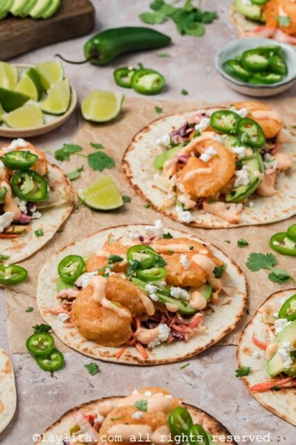 Baja shrimp tacos on brown parchment paper topped with sauce and sliced jalapeños with lime slices on the side