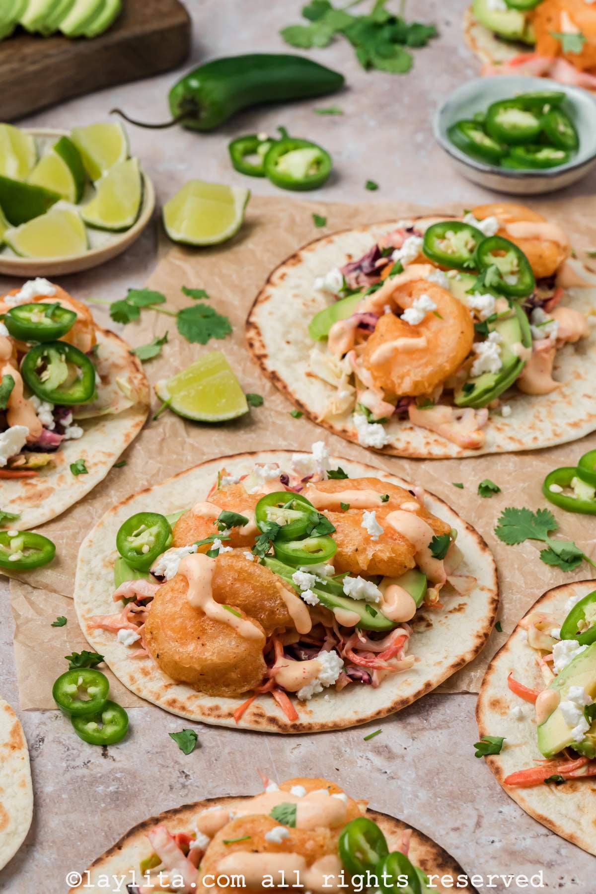 Baja shrimp tacos on brown parchment paper topped with sauce and sliced jalapeños with lime slices on the side