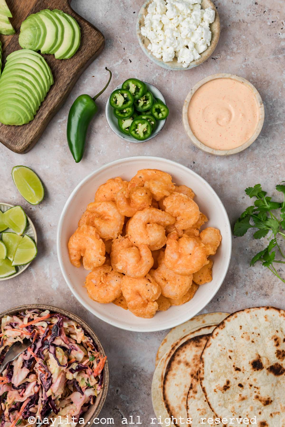 Overhead view of a plate of beer battered fried shrimp next to crispy tortillas, cabbage slaw, baja tacos slices, sliced avocados, jalapeños and lime slices