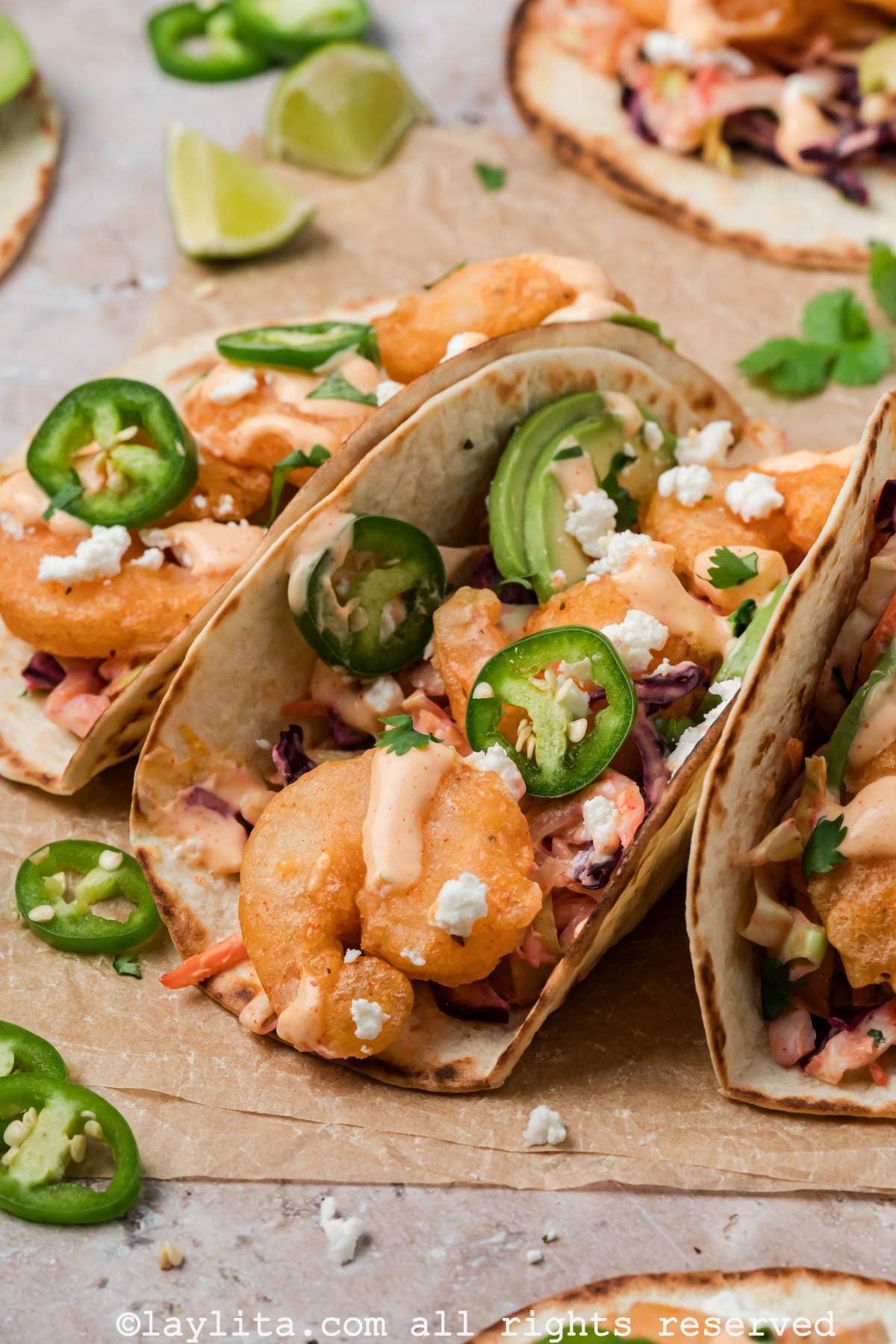 Side view of three shrimp tacos stacked against each other on a piece of parchment paper, topped with sauce, jalapeños and avocado slices