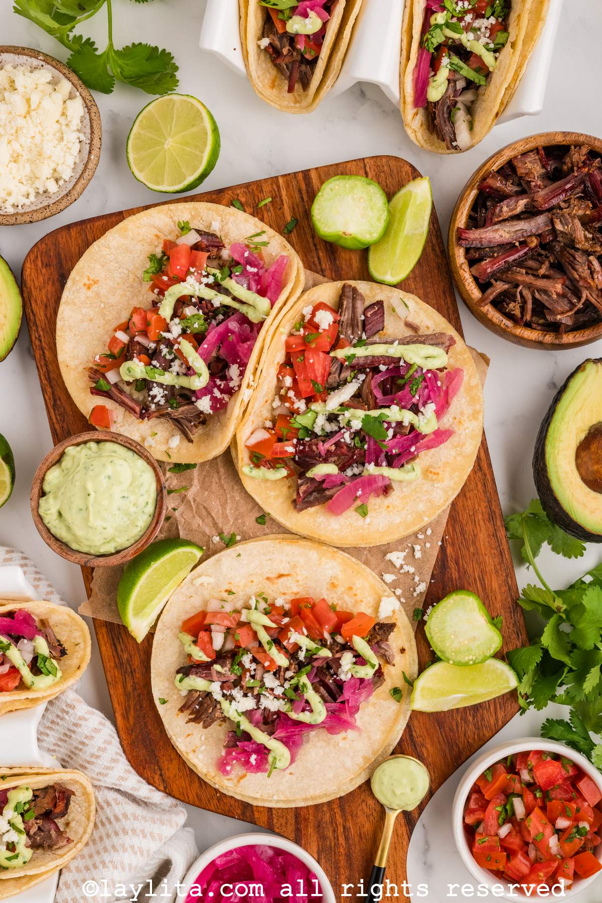 Overhead shot of three beef brisket tacos on a wooden board next to sauces, lime wedges and cilantro.