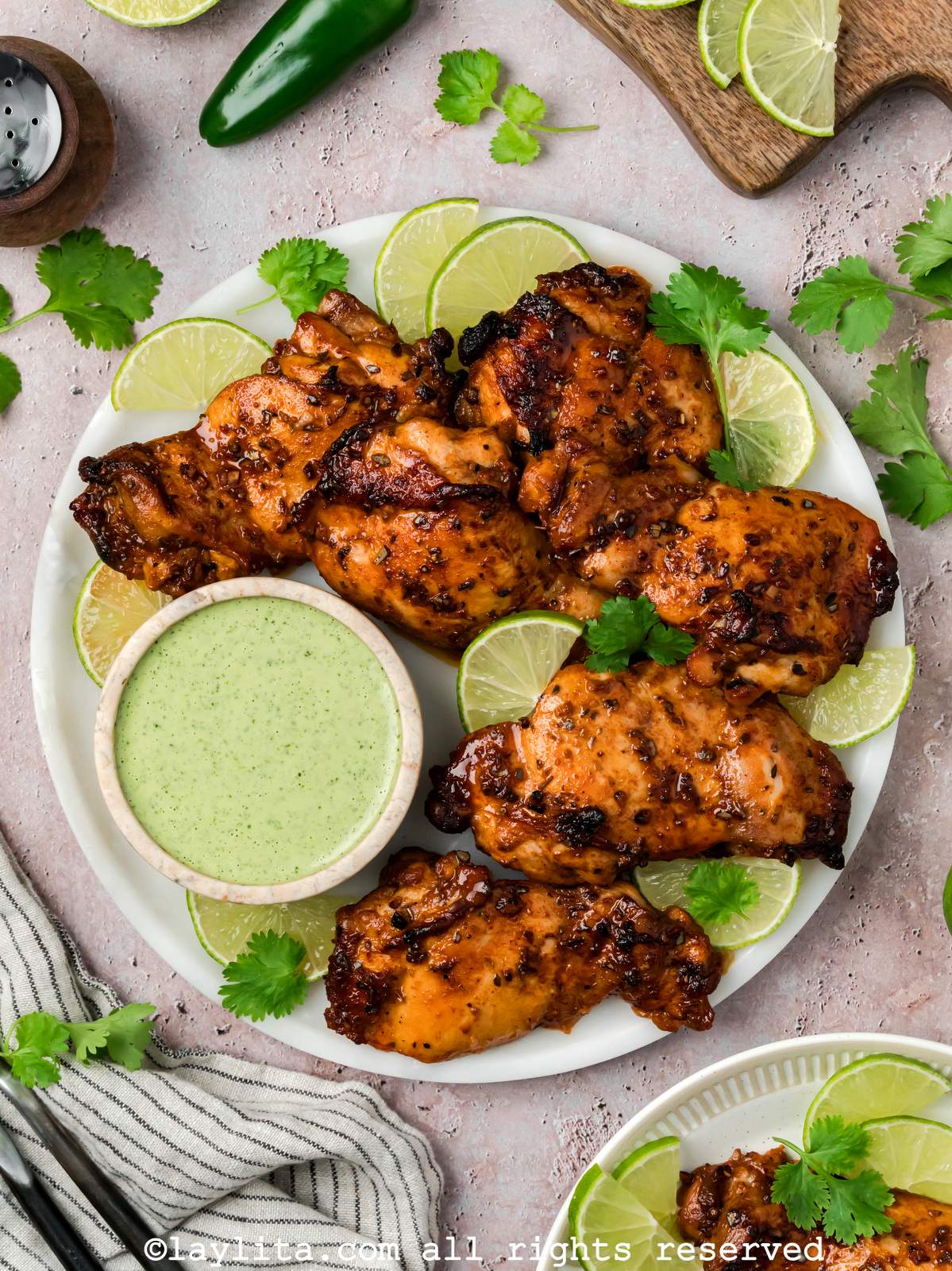 Overhead shot of Peruvian chicken thighs on a plate next to a small bowl of green sauce. With cilantro leaves, lime slices and jalapeños on the side of the plate.