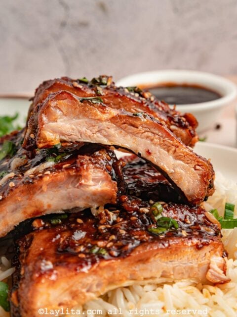 Closeup view of several sticky pork ribs sliced into pieces and stacked on top of each other on top of rice, with a bowl of sauce in the background
