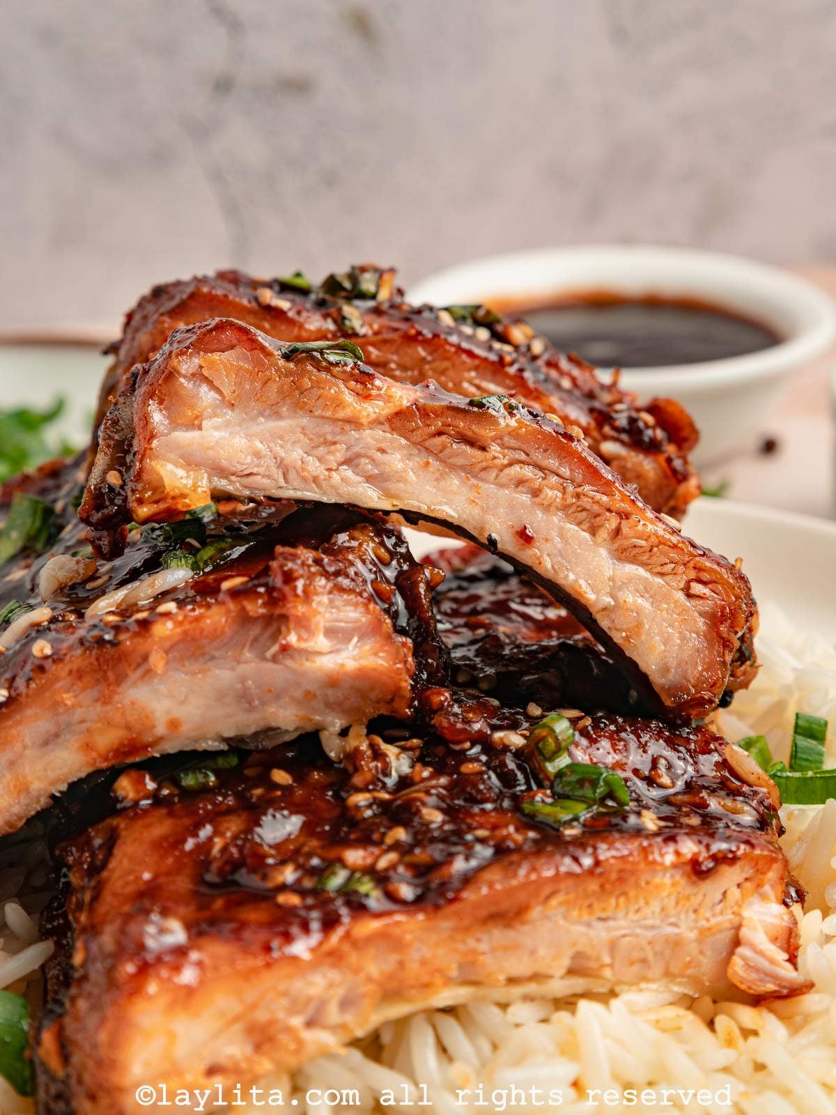 Closeup view of several sticky pork ribs sliced into pieces and stacked on top of each other on top of rice, with a bowl of sauce in the background