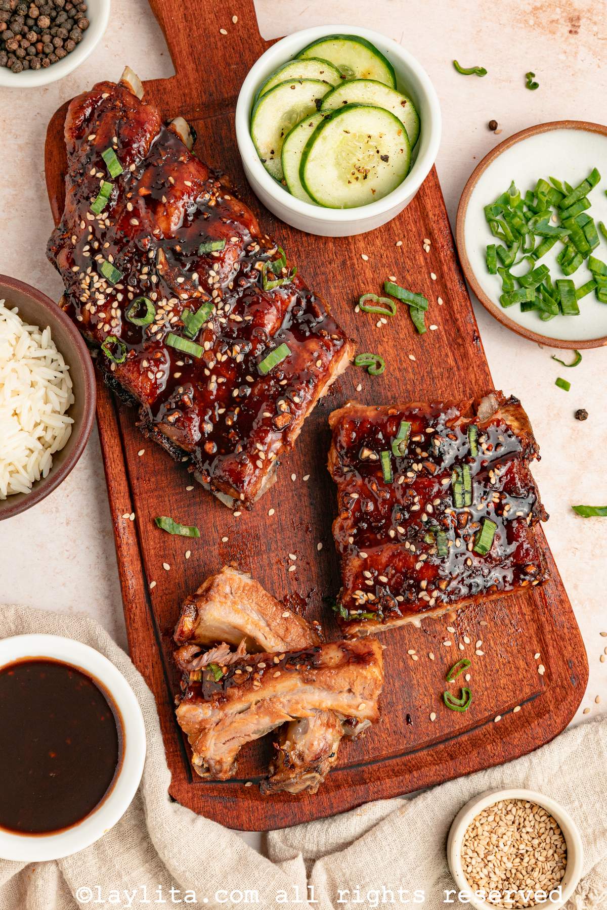 Overhead view of Asian style ribs on a board, with some cut into slices to show the meat, surrounded by small bowls of cucumber salad, rice, sauce and herbs