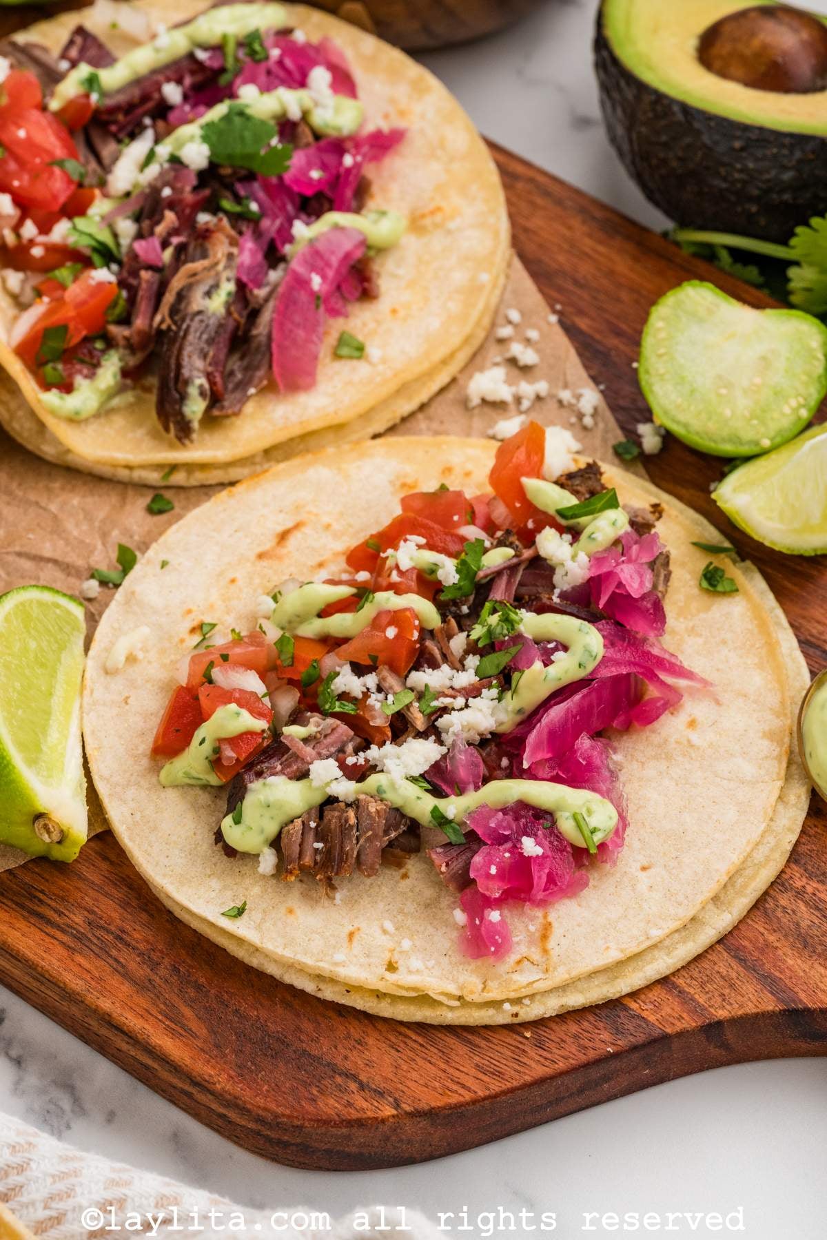 Two beef brisket tacos on a wooden board, topped with pickled onions, pico de gallo and green sauce.