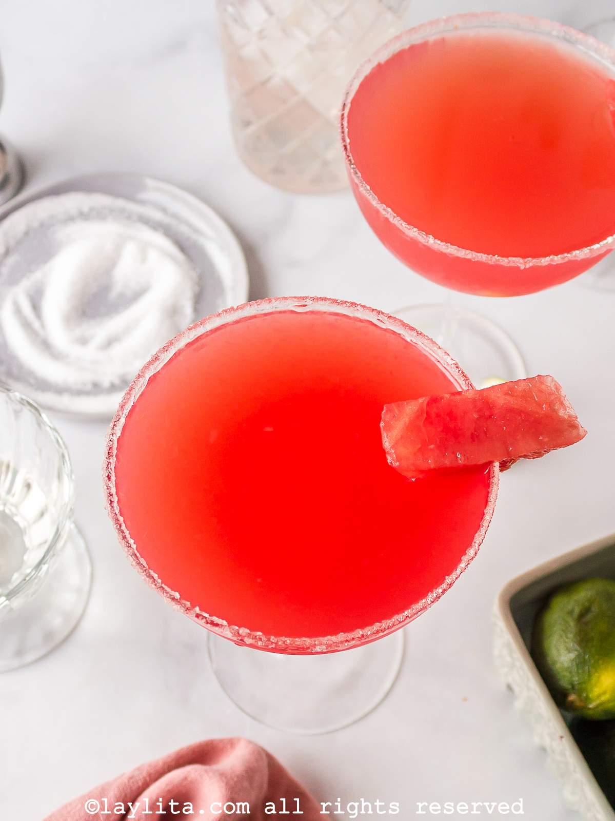 Close up of a watermelon margarita in a coupe glass garnished with sugar and a piece of watermelon.