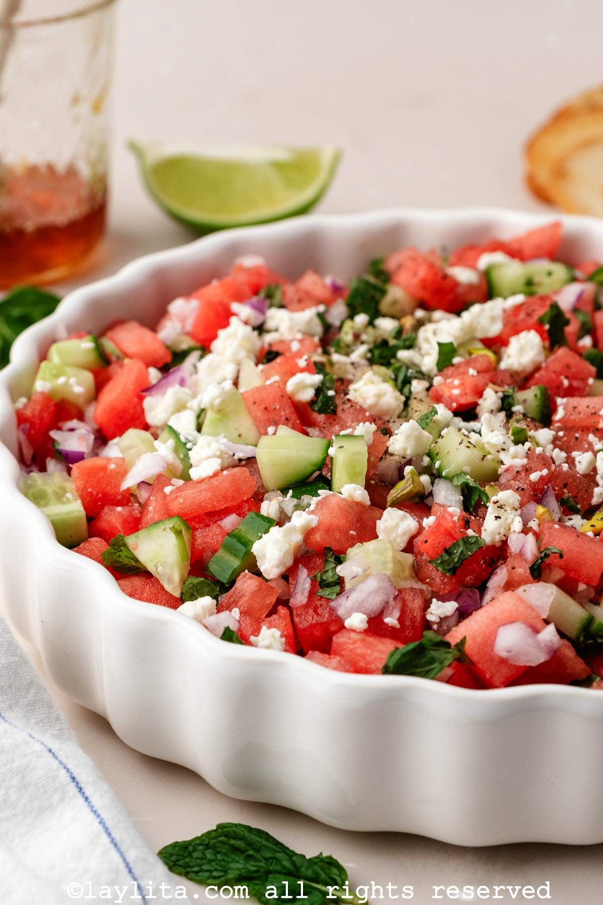 Side view of a bowl of watermelon cucumber feta salad with a lime wedge and slice of bread in the background