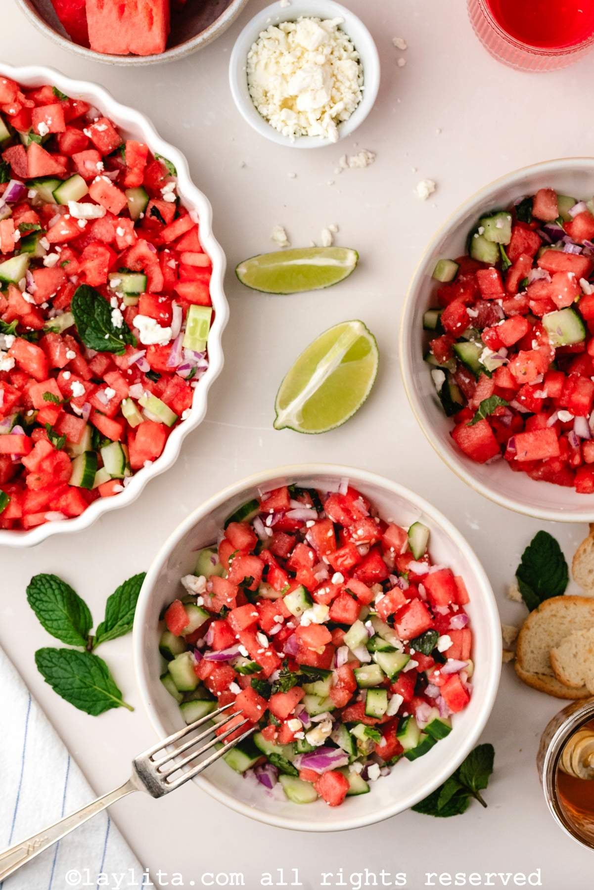Overhead view of three bowls of watermelon salad with lime wedges, mint leaves and crumbled feta cheese on the side