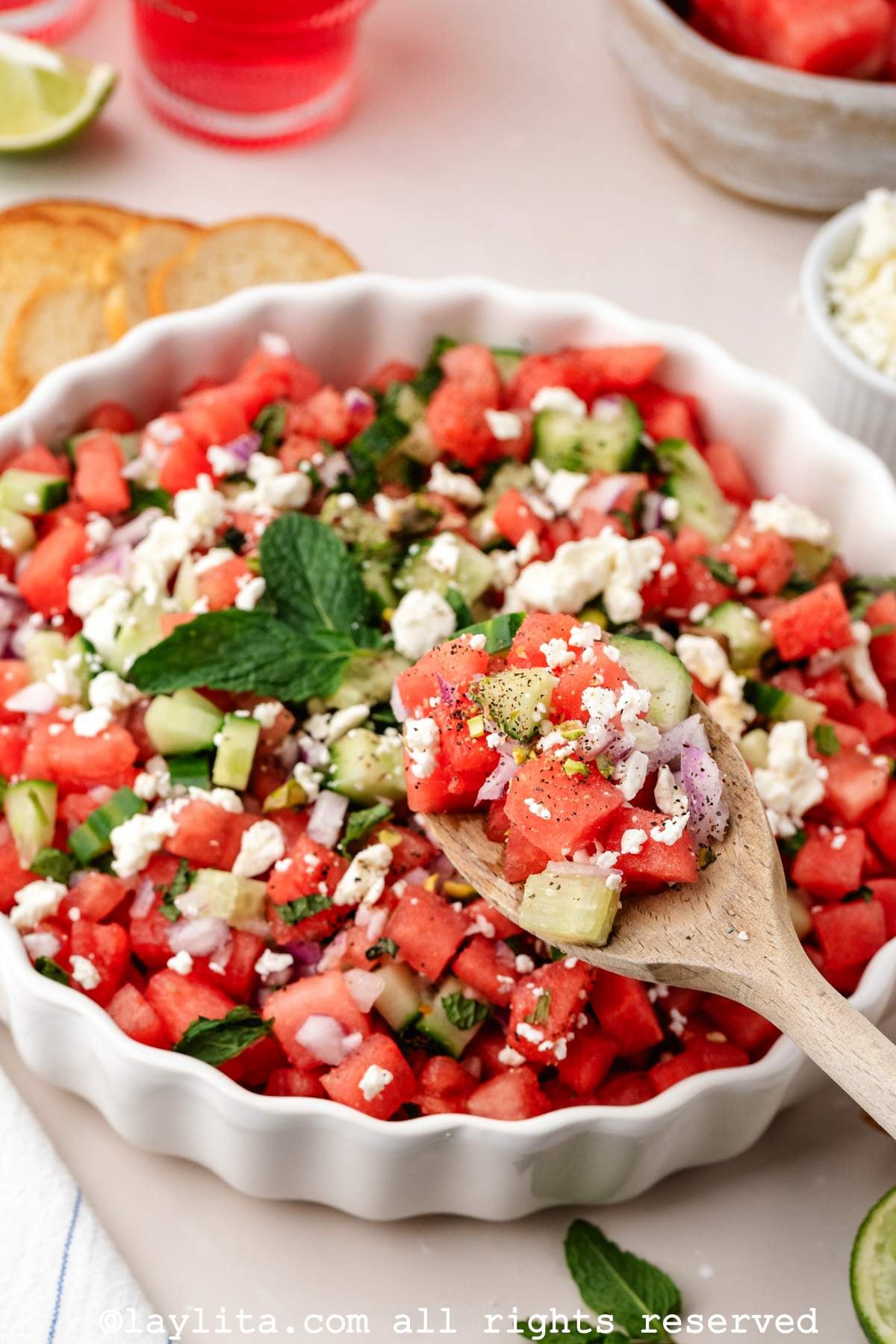 A bowl of watermelon feta salad topped with mint leaves, with a serving spoon scooping some salad out of the bowl