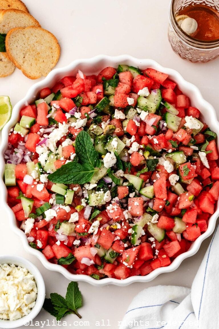 Overhead view of a watermelon feta salad in a white serving bowl, around the bowl there are pita chips, honey, mint leaves and a bowl of crumbled feta cheese