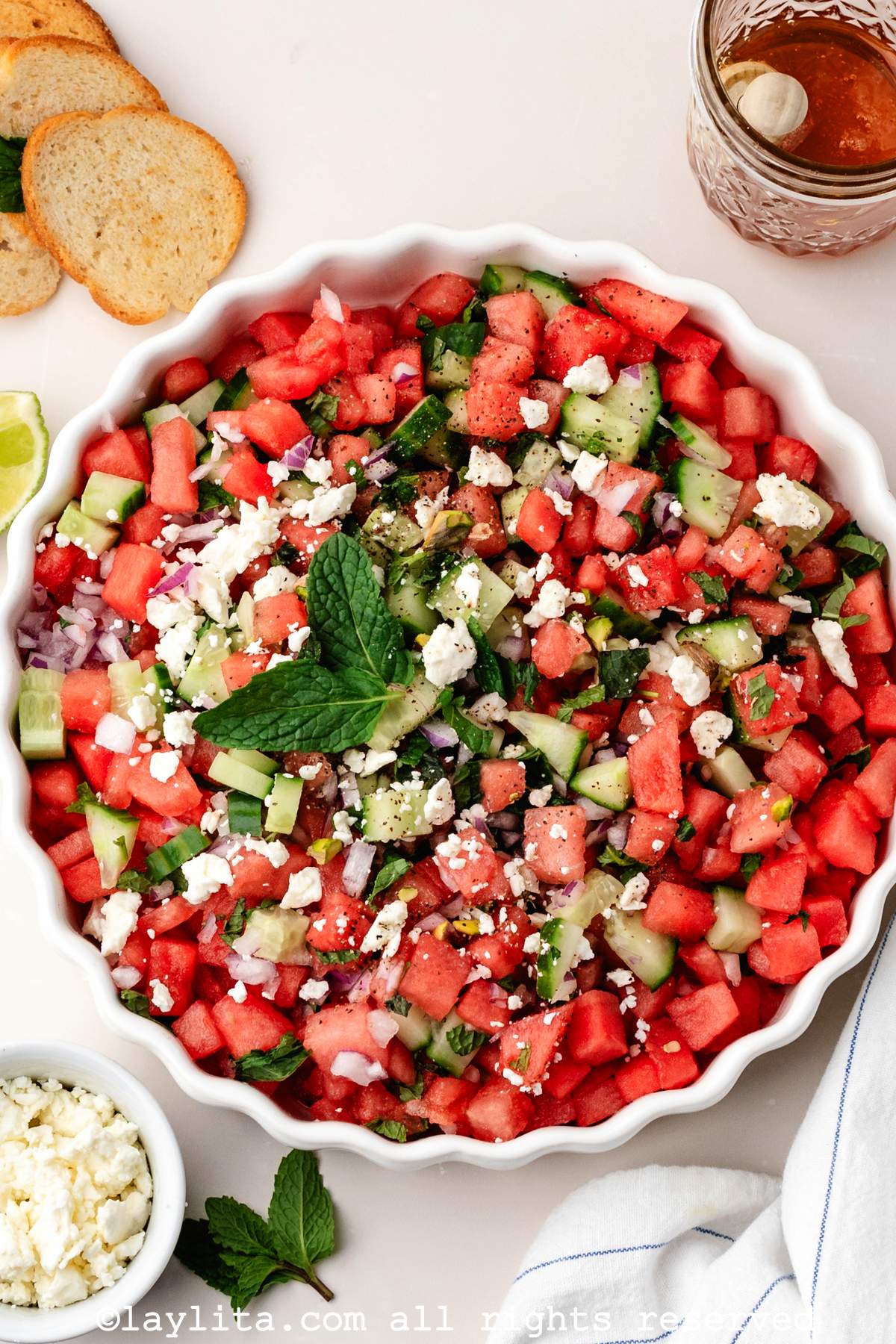 Overhead view of a watermelon feta salad in a white serving bowl, around the bowl there are pita chips, honey, mint leaves and a bowl of crumbled feta cheese