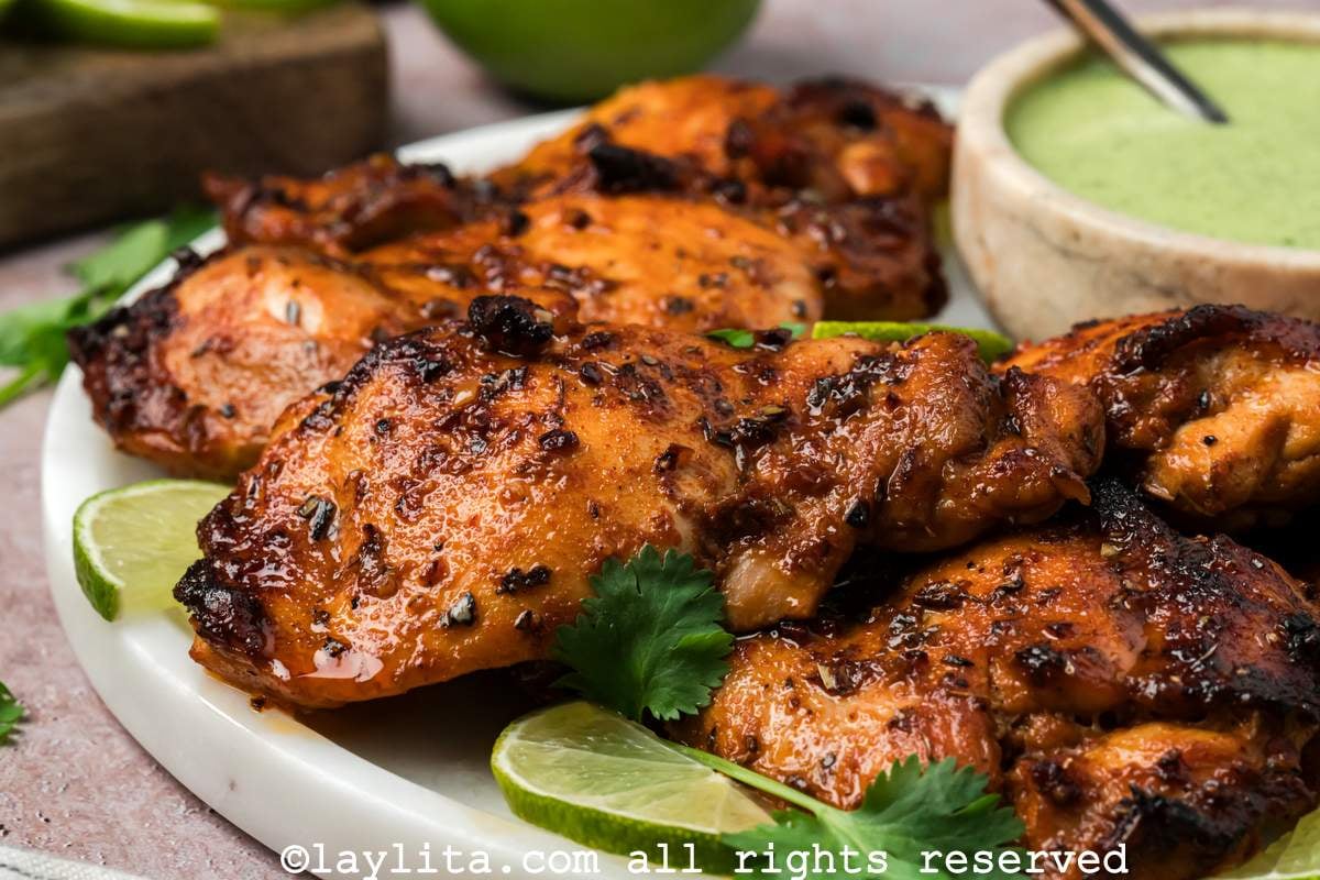 Side view of a plate with roasted chicken thighs, served with lime slices and a bowl of green sauce