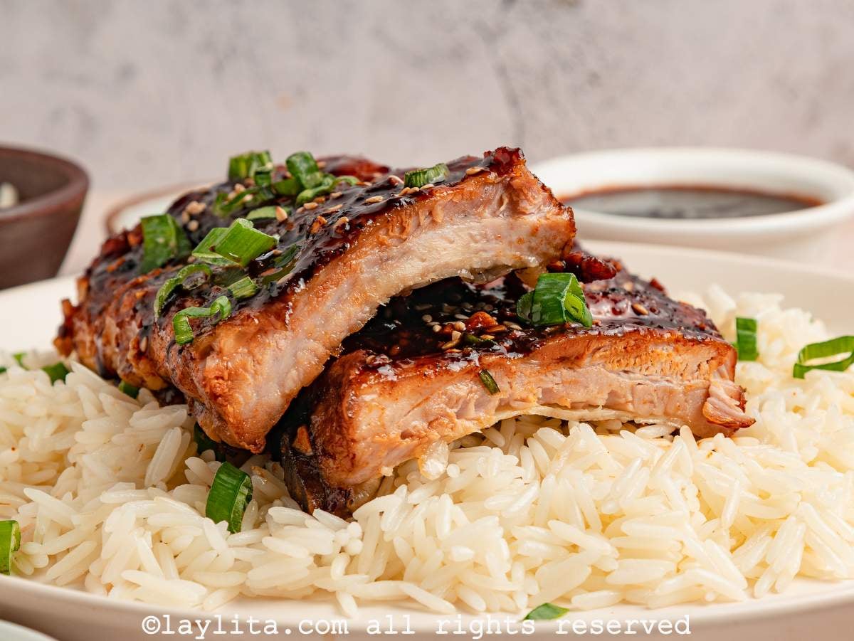 Sticky pork ribs sliced into pieces and stacked on top of each other and garnished with green onions in a bowl of rice, with a bowl of sauce in the background