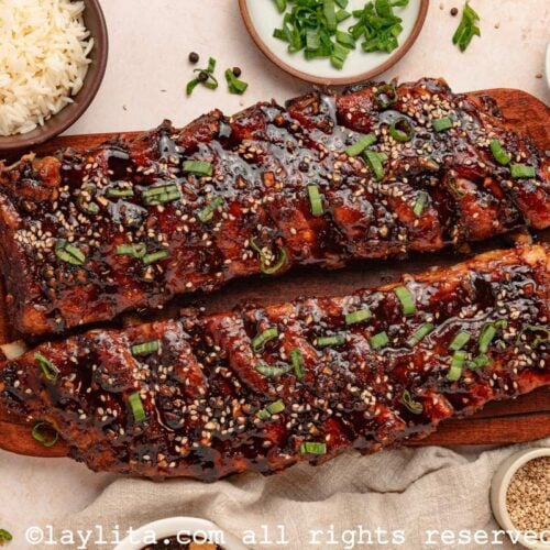 Overhead shot of two racks of Asian sticky ribs on a wood board surrounded by bowls of rice, cucumber, sesame seeds, and herbs