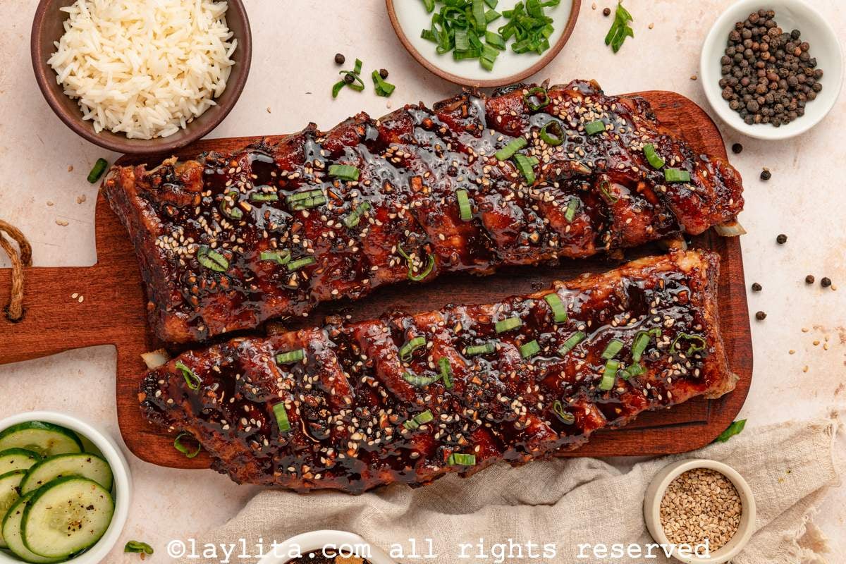 Overhead shot of two racks of Asian sticky ribs on a wood board surrounded by bowls of rice, cucumber, sesame seeds, and herbs