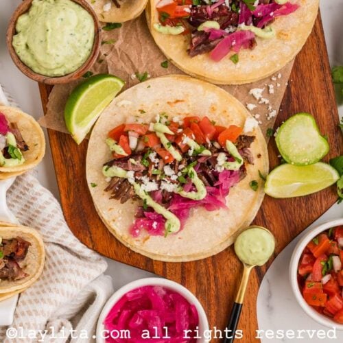 Overhead shot of several shredded beef brisket tacos with green salsa, pico de gallo, pickled onion, cilantro and lime wedges on the side