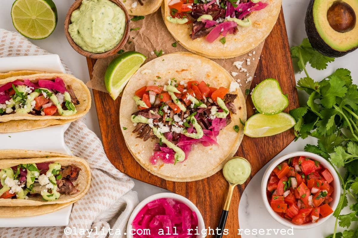 Overhead shot of several shredded beef brisket tacos with green salsa, pico de gallo, pickled onion, cilantro and lime wedges on the side