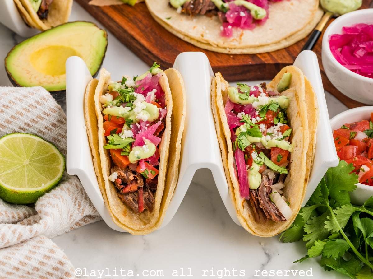 Two brisket tacos in a taco holder with green sauce, pico de gallo, pickled onions and cheese crumbles on top
