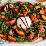 Overhead view of a salad made with grilled peach wedges, cherry tomato, burrata cheese, arugula, basil leaves and balsamic glaze on an oval platter