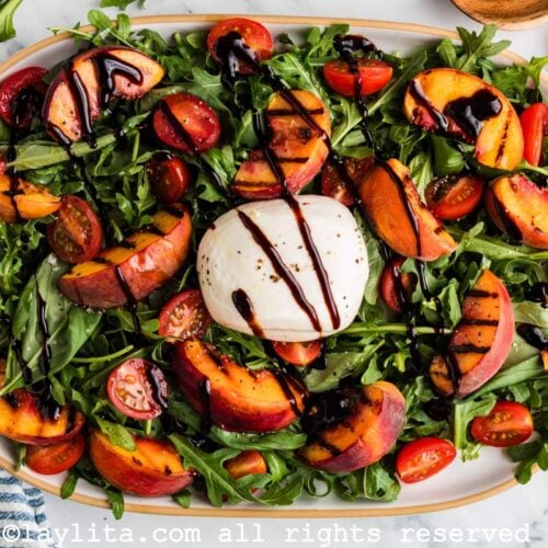 Overhead view of a salad made with grilled peach wedges, cherry tomato, burrata cheese, arugula, basil leaves and balsamic glaze on an oval platter