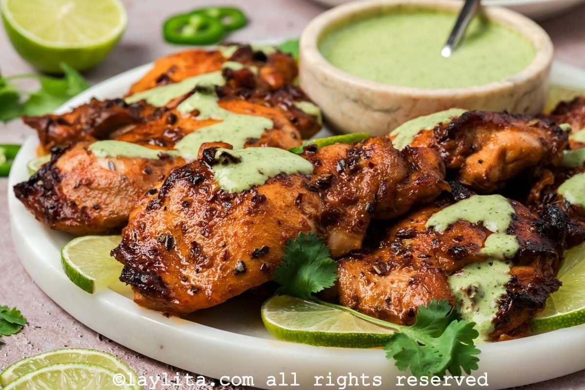 Side view of Peruvian chicken thighs on a plate next to a small bowl of green sauce or ají verde.