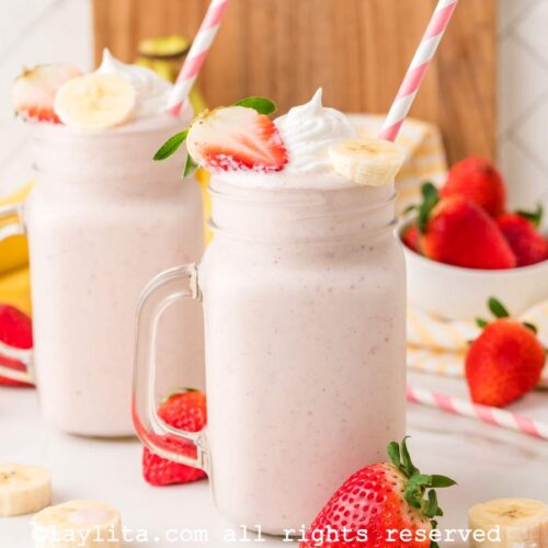 Side view of two large jars of strawberry banana milkshakes with fresh strawberries and bananas scattered around.