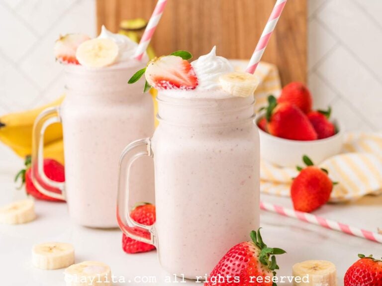 Side view of two large jars of strawberry banana milkshakes with fresh strawberries and bananas scattered around.