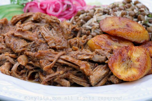 Closeup of juicy pulled pork on a plate with fried ripe plantains, lentils, rice and pickled onions